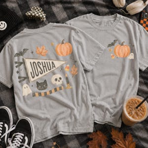 Puede incluir: Dos camisetas gris claro con gráficos de Halloween. El diseño trasero muestra un banderín con el nombre JOSHUA, una calabaza, un gato negro, una calavera, un fantasma, una vela y hojas. El diseño frontal muestra una calabaza y un fantasma.