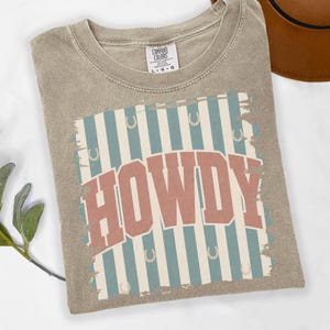 Peut inclure: T-shirt Comfort Colors beige avec l'inscription "HOWDY" dans une police r&eacute;tro. Le motif pr&eacute;sente des rayures bleues et blanches avec des fers &agrave; cheval. Le t-shirt est pli&eacute; sur une surface blanche.