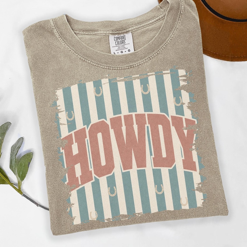 Peut inclure: T-shirt Comfort Colors beige avec l'inscription "HOWDY" dans une police r&eacute;tro. Le motif pr&eacute;sente des rayures bleues et blanches avec des fers &agrave; cheval. Le t-shirt est pli&eacute; sur une surface blanche.