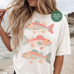 Puede incluir: Camiseta color crema con tres ilustraciones estilizadas de peces en tonos rojos, melocotón y verde. Los peces están dispuestos verticalmente, con bocas abiertas y aletas detalladas. La camiseta tiene un corte holgado.