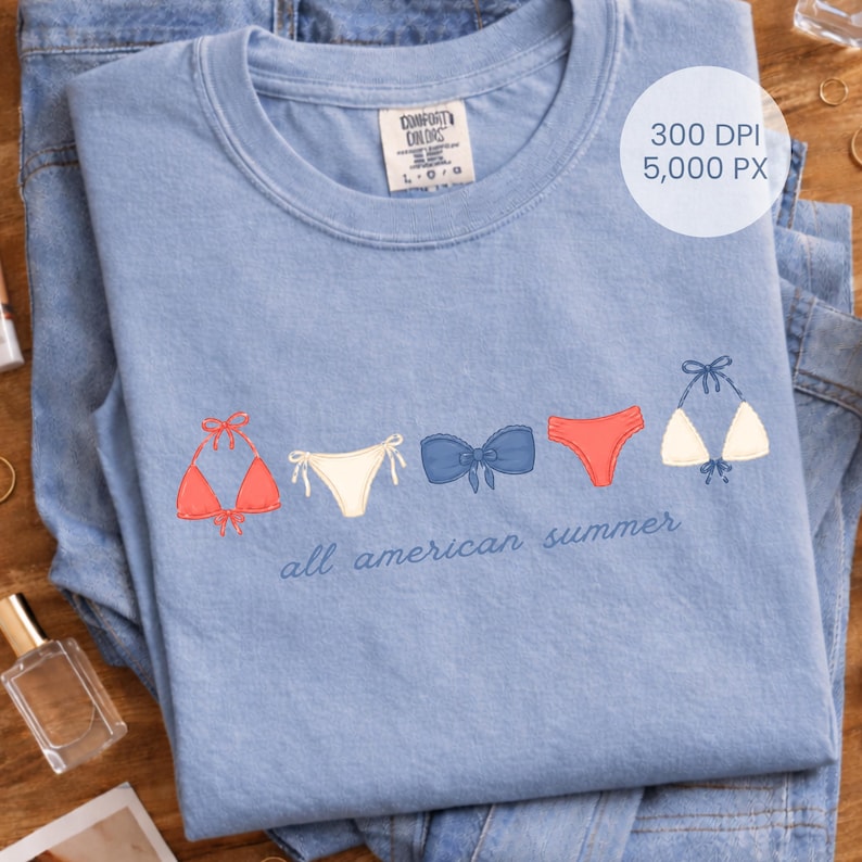 Peut inclure: T-shirt bleu clair avec des motifs de bikinis brod&eacute;s en rouge, blanc et bleu. L'inscription "all american summer" est &eacute;crite en cursive en dessous. Le t-shirt est pli&eacute; sur un jean bleu.