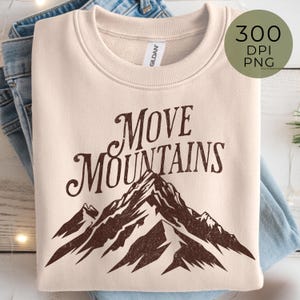 May include: A cream-colored sweatshirt with the text "MOVE MOUNTAINS" in a brown, vintage-style font. Below the text is a brown mountain graphic. The sweatshirt is folded on top of a pair of blue jeans.