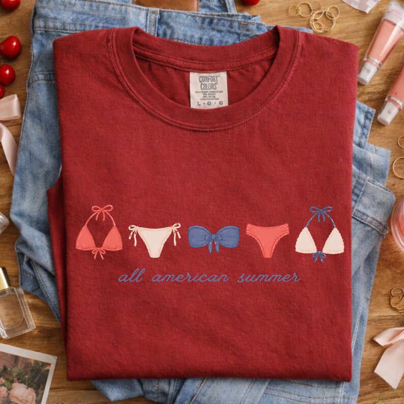 Peut inclure: Un t-shirt Comfort Colors bordeaux avec des motifs de bikinis brod&eacute;s en corail, blanc et bleu. L'expression "all american summer" est brod&eacute;e en dessous. Le t-shirt est pli&eacute; sur un jean bleu.