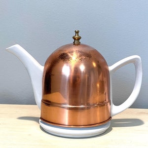 May include: A white teapot with a copper lid and a brass knob. The teapot has a white base and a white handle.