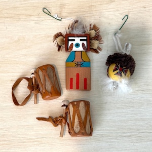 May include: A collection of Native American-style ornaments and miniature drums. The ornaments feature painted wooden figures with feather accents. The drums are made of leather and wood, with leather straps. The color palette includes brown, red, blue, and white.