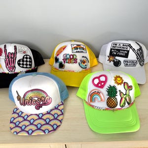 Peut inclure: Cinq casquettes de camionneur colorées avec divers patchs et broderies. Les casquettes présentent des motifs comme une licorne, de bonnes vibrations, un signe de paix, un ananas et une main de squelette.