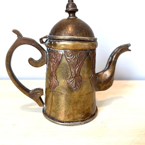 May include: Antique brass teapot with a curved handle and spout. The body features ornate copper-colored panels with a floral design. The lid is domed and topped with a small finial. The teapot has a vintage aesthetic.