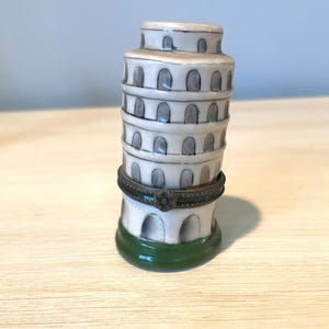 May include: A small ceramic trinket box shaped like the Leaning Tower of Pisa. The box is white with gray arches and a green base. It has a gold-colored metal hinge.