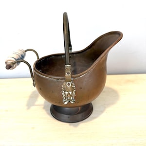 May include: A copper-colored coal scuttle with a handle and a decorative handle on the side. The scuttle has a round base and a spout. The handle is made of metal and the side handle is white and blue. The scuttle is likely antique.