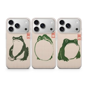 May include: Three clear phone cases featuring green ink-style frog illustrations. The first shows a frog with a bowl, the second a round frog, and the third a seated frog. Each case includes a small red rectangular stamp with Japanese text.