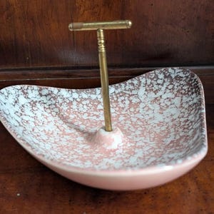 May include: A kidney-shaped dish featuring a pink and white speckled pattern with a central vertical brass-colored metal handle. The dish has a smooth, curved rim and a solid pink base, with the metal handle extending upward from the center.