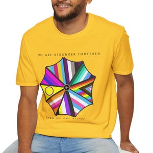 May include: A yellow t-shirt with a colorful umbrella graphic featuring various pride flags. The text "WE ARE STRONGER TOGETHER" is above the graphic, and "THAN WE ARE ALONE" is below. The shirt is made of a soft material.