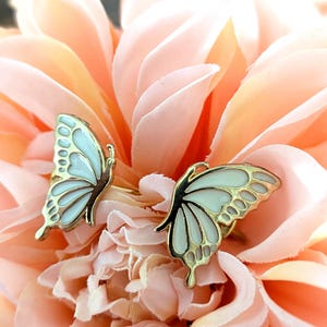 May include: Two butterfly-shaped rings with gold-colored metal and white and light blue enamel wings. The rings are set against a soft, peach-colored floral background, creating a delicate and feminine aesthetic. The butterfly wings have detailed patterns.