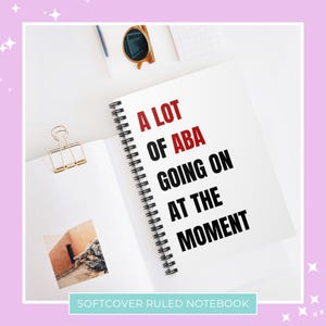 以下が含まれることがあります： 黒いスパイラルバインディングが付いた白いノート。表紙には「A LOT OF ABA GOING ON AT THE MOMENT」というテキストが黒と赤の文字で書かれています。ノートには「Softcover Ruled Notebook」というラベルが貼られています。