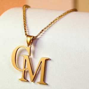 Puede incluir: Un collar dorado con un colgante con las letras "GM". El colgante está conectado a una delicada cadena. El collar se muestra sobre un fondo blanco.