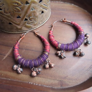 May include: A pair of hoop earrings with a unique design. The hoops are wrapped in red and purple fabric and adorned with small, dangling metal charms. The earrings have a bohemian or ethnic aesthetic.