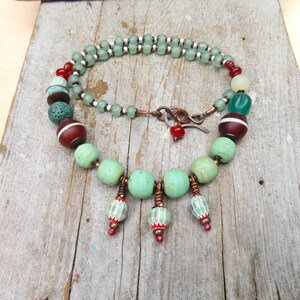 以下が含まれることがあります： 緑、赤、茶色のビーズを使った手作りのネックレス。 ネックレスは銅製の留め金と、装飾用のぶら下がりビーズが付いています。 ビーズは繊細なチェーンで繋がれています。 ユニークなジュエリーです。