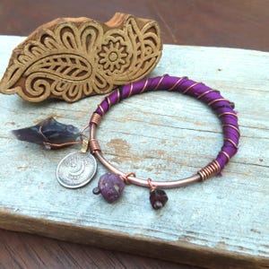May include: A handmade bracelet with a copper band wrapped in purple fabric and copper wire. The bracelet features a silver coin charm, a dark purple stone, and a small, dark red stone. A wooden block with a carved floral design is in the background.