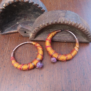 Puede incluir: Un par de pendientes de aro. Cada pendiente presenta un marco de alambre de cobre envuelto en tela naranja y roja. Una pequeña gema púrpura está unida a la parte inferior de cada pendiente. Los pendientes se exhiben sobre una superficie de madera.