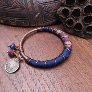May include: A copper bangle bracelet with a blue fabric wrap and copper wire accents. The bracelet features a small coin charm and dangling beads in shades of blue and red. The bracelet is on a wooden surface.