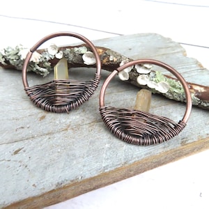 May include: A pair of copper wire wrapped hoops with clear quartz crystals inside. The hoops are on a wooden surface.