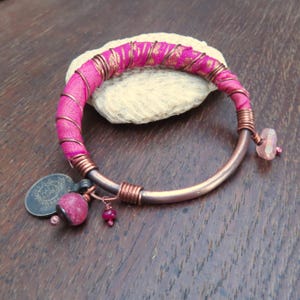 May include: A handcrafted copper bracelet with a pink fabric wrap and gold detailing. The bracelet includes copper wire, a pink gemstone, and a small coin charm. Small red gemstones are also attached to the bracelet.