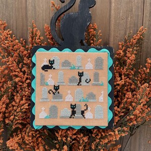 以下が含まれることがあります： 黒猫と墓石がベージュの背景に描かれたクロスステッチのデザイン。ターコイズのスカラップエッジで縁取られています。黒猫のシルエットがフレームの上にあります。オレンジ色の装飾的な葉で囲まれ、ハロウィーンをテーマにしています。