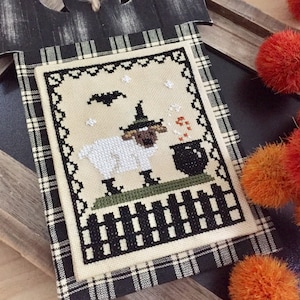 May include: A white sheep wearing a witch's hat and holding a candy cane stands in front of a black cauldron with green steam coming out of it. The sheep is on a green patch of grass with a black and white picket fence behind it. The image is framed with a black and white plaid border.