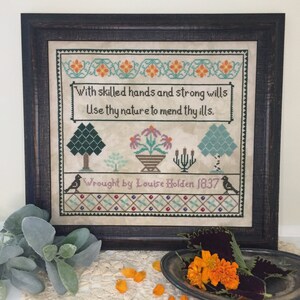 Pode incluir: Um sampler de ponto cruz emoldurado com uma citação sobre o uso da natureza para curar doenças. O sampler está em uma moldura de madeira escura e apresenta um design floral com pássaros e uma cesta de flores. O texto diz "With skilled hands and strong wills Use thy nature to mend thy ills. Wrought by Louise Holden 1837".