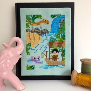 May include: A cross-stitch pattern featuring a jungle scene with a waterfall, a river, a frog, a snake, and a person in a boat. The scene is framed in a black frame.