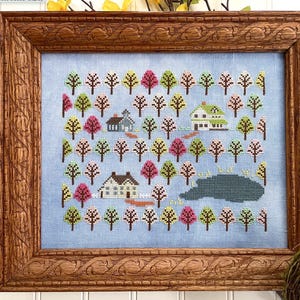 Könnte beinhalten: Gerahmte Kreuzstichlandschaft mit Bäumen, Häusern und einem See. Die Bäume haben rosa, grüne und gelbe Blätter. Die Häuser sind weiß und blau. Der Rahmen ist aus braunem Holz.