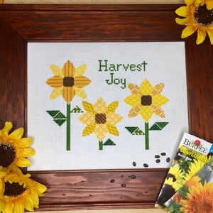 May include: A framed cross-stitch embroidery featuring three yellow sunflowers and the text "Harvest Joy". The embroidery is surrounded by a dark wood frame. Two packets of sunflower seeds are visible in the bottom right corner of the image.