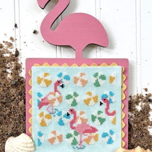 May include: A pink flamingo-shaped wooden cutout above a cross-stitch design. The cross-stitch features pink flamingos, beach balls, and green foliage on a light blue background. The design is framed with a pink border and yellow trim.
