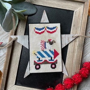 May include: A cross-stitch design featuring a white dog wearing a red, white and blue hat and riding a red, white and blue firework rocket. The design is framed in a wooden frame with a star-shaped cutout.