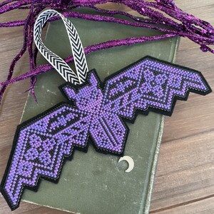 以下が含まれることがあります： 黒と紫の刺繍が施されたコウモリのオーナメントで、黒と白のリボンループが付いています。コウモリは、銀色の三日月がついた緑色の本にぶら下がっています。