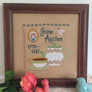 Op de afbeelding: Een ingelijst borduurwerk met kruissteek, met een portret van Jane Austen, een theepot, een theekopje en bloemen. De tekst luidt "Jane Austen 1775-1817".