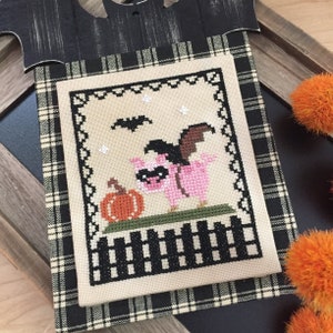May include: A cross-stitch pattern of a pink pig wearing a black and brown bat costume, holding a pumpkin. The pig is standing on a green patch of grass with a black and white picket fence below. The background is a cream color with a few white stars and a black bat flying in the sky.