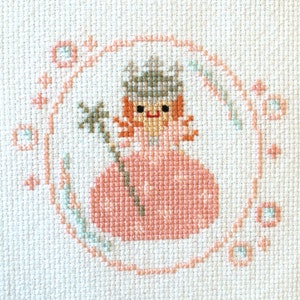 May include: A cross-stitch embroidery of a girl wearing a pink dress and a gray hat. The girl is holding a flower and is surrounded by a pink and green floral design.