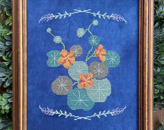 Puede incluir: Obra de arte en punto de cruz enmarcada con un diseño floral. La obra presenta flores naranjas y marrones, junto con hojas verdes y marrones, sobre un fondo azul oscuro. El marco es de color marrón dorado.