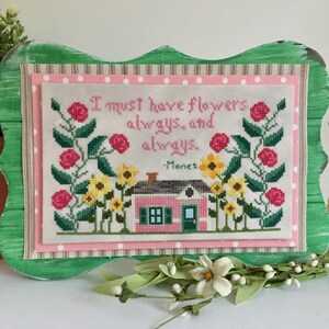 May include: Cross-stitch embroidery of a pink house with a green door, surrounded by sunflowers and pink roses. The text reads "I must have flowers always and always - Monet".