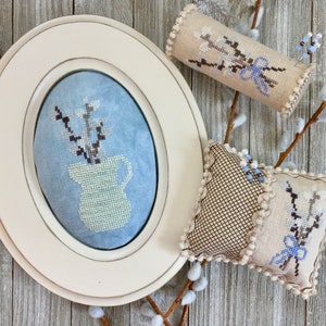 May include: An oval, off-white framed cross-stitch featuring a light blue background with a white pitcher holding branches. Two decorative pillows and a cylindrical pin cushion, all with matching floral embroidery and light blue bows, are also present.