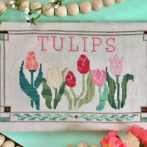 May include: A cross-stitch embroidery of a floral design featuring a bouquet of pink, red, and white tulips with green leaves. The embroidery is framed with a brown and green border and the word "TULIPS" is stitched in pink above the flowers.
