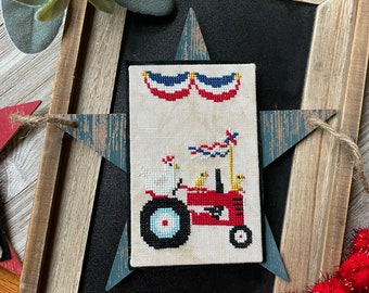 May include: A cross-stitch design featuring a red tractor with a white and blue flag, pulling a wagon with three chicks. The tractor is decorated with red, white, and blue bunting.