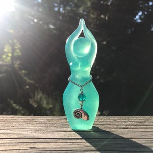 Puede incluir: Una estatua de diosa verde azulada con los brazos levantados, un collar de cadena plateada con un cristal verde azulado y un colgante de concha plateada.