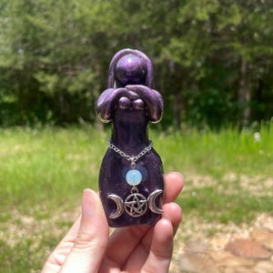 May include: A purple ceramic goddess figurine with a silver chain necklace and a pendant featuring a light blue stone and a triple moon pentacle charm. The figurine has a glossy finish and is held against a blurred green background.