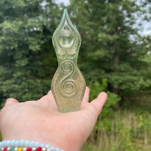 May include: A translucent green resin goddess figurine with glitter accents. The sculpture has a pointed top and a spiral design on the torso. The figurine is held in a hand against a blurred green background.