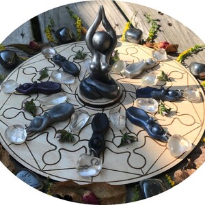May include: A wooden circle with a geometric design and a black figurine of a woman in a yoga pose in the centre. The circle is surrounded by smaller black and clear figurines and stones.