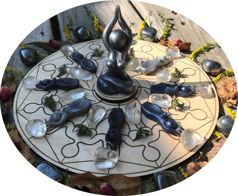 May include: A wooden circle with a geometric design and a black figurine of a woman in a yoga pose in the centre. The circle is surrounded by smaller black and clear figurines and stones.