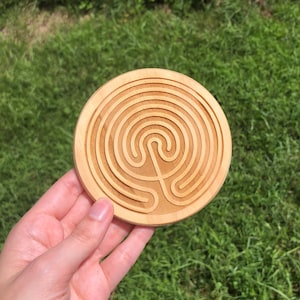 Puede incluir: Un posavasos redondo de madera clara con un diseño de laberinto tallado. El posavasos se sostiene en una mano sobre un fondo de hierba verde. El patrón del laberinto presenta círculos concéntricos y un camino central.