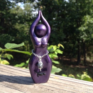 May include: A purple ceramic figurine of a woman in a yoga pose with a silver chain and a black stone pendant.